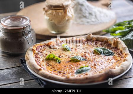 Pizza Napoli with tomato sauce, mozzarella and fresh basil, in the background flour and a jar of rye sourdough. Traditional Italian pizza with sourdou Stock Photo
