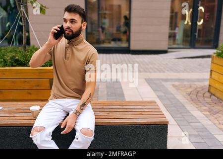 Portrait of confident tattooed young man talking on mobile phone sitting on bench on city street. Bearded male having conversation on smartphone Stock Photo