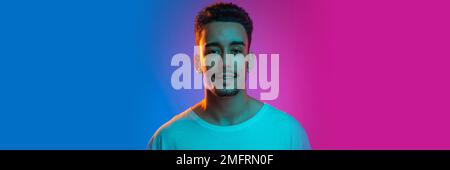 Positivity. Young man in casual white T-shirt posing, looking at camera over gradient blue pink background in neon light. Banner, flyer Stock Photo