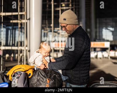 Fatherat comforting his tired infant baby boy child sitting on top of luggage cart in front of airport terminal station while traveling wih family Stock Photo