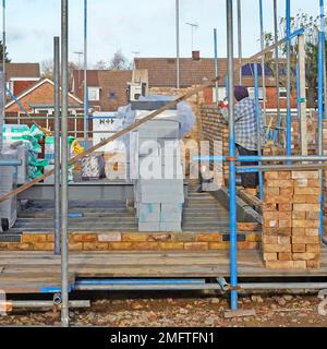 Bricklayer working amongst scaffolding & building construction materials early stages of brick cavity wall structure of new detached house England UK Stock Photo