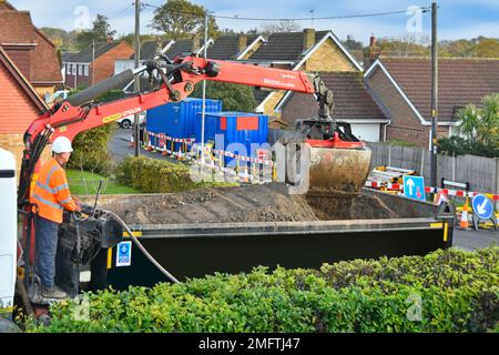 Tipper truck driver standing operating controls grab crane bucket loader on back of dual purpose lorry beside road works in residential England UK Stock Photo
