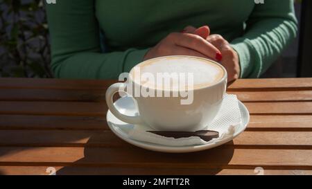 Girl at the table in a cafe with a cup of cappuccino. Delicious drink with appetizing milk foam. Cup of coffee close-up. Breakfast outdoors. Stock Photo