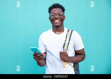 Portrait of happy young man smiling with a mobile phone and a backpack on turquoise background - Portrait of a cheerful young man - Happines and Stock Photo