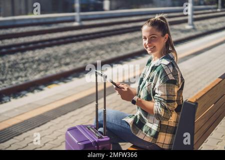 Happy woman using mobile phone while sitting on a bench at the train station. Stock Photo