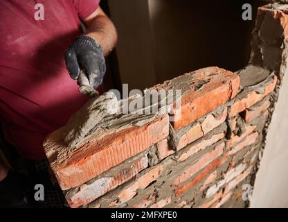 Close up of male hand in work glove laying brickwork in building under construction. Man bricklayer builder applying cement mortar on bricks with trowel tool. Masonry construction concept. Stock Photo