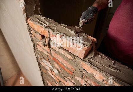 Close up of man hand in work glove laying brickwork in building under construction. Male worker applying cement mortar on bricks with trowel tool. Masonry construction concept. Stock Photo