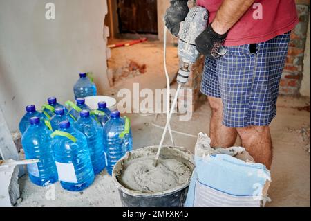 Close up of man builder in work gloves preparing sand-cement mix for bricklaying in building under construction. Male worker mixing mortar with electric cement concrete mixer. Stock Photo