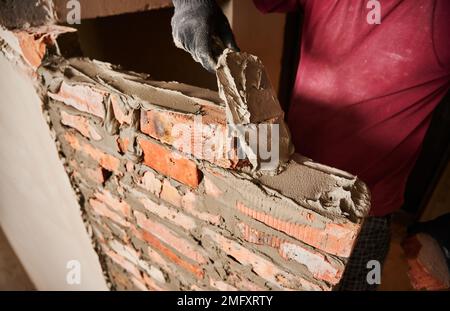 Close up of man hand in work glove laying brickwork in building under construction. Male worker applying cement mortar on bricks with trowel tool. Masonry construction concept. Stock Photo
