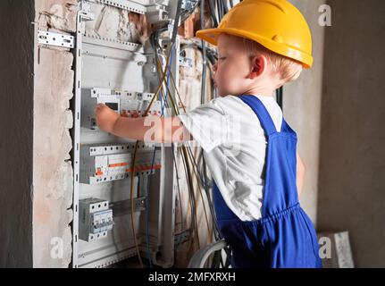 Side view of child in safety helmet fixing electrical switchboard or distribution board in apartment under renovation. Baby electrician repairing household electrical panel with automatic fuses. Stock Photo