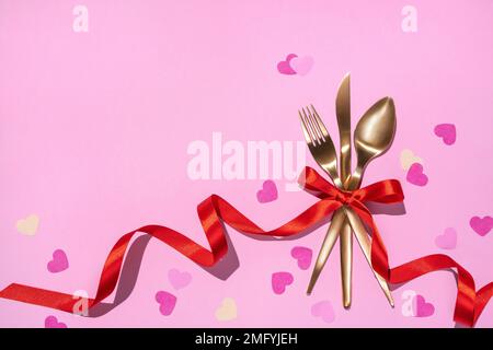 Golden fork, knife, spoon tied with red ribbon on pink background. Valentine's Day or festive romantic dinner. Stock Photo