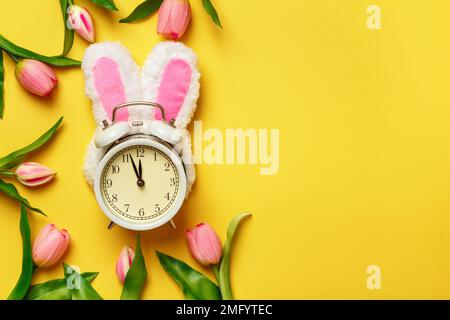 Alarm clock in rabbit ears surrounded by tulips. Easter spring change of seasons. Stock Photo