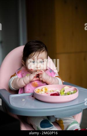 Cute baby girl sitting on highchair in kitchen, eating raw vegetables, fruits and smiling. Healthy eating for baby concept. Mom's daily lifestyle. Stock Photo