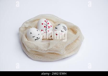 Three beautifully painted Easter eggs lie in environmentally friendly mesh bag on white table. Funny faces and twig with red flowers are painted on eggs. Copy space. Happy holidays Stock Photo