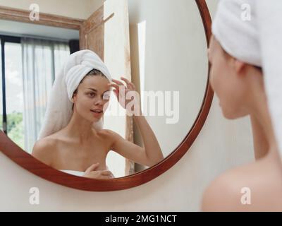 A beauty woman stands in front of a mirror after a shower in a towel on her head looks at her reflection and does a facial massage applies a day cream Stock Photo