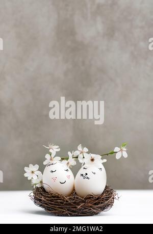 Happy Easter. White Easter eggs in wicker wooden nest on white table on gray background. View is straight. Blossoming branch of cherry with white flowers. Stylish spring template with copy space Stock Photo