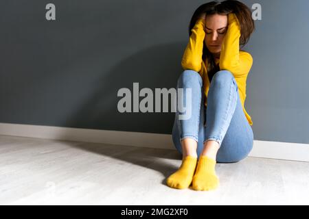 Full length of sad suffering young female in yellow sweater and socks touching head while sitting on floor near gray wall with closed eyes Stock Photo