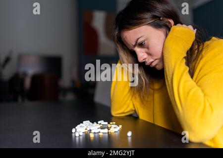 Side view of melancholic serious female with long hair in yellow sweater sitting at table and looking at pile of antidepressant pills at home Stock Photo