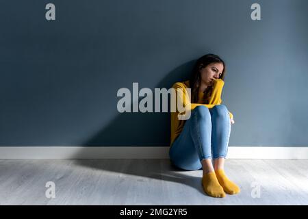 Full body of young melancholic lady with long dark hair in jeans yellow sweater and socks sitting on parquet floor near gray wall  Stock Photo
