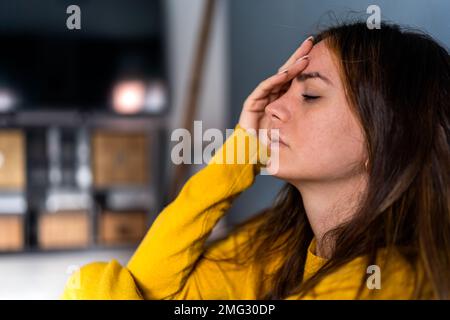 Side view of unhappy young female with long dark hair in casual clothes sitting on floor with closed eyes and touching forehead at home Stock Photo