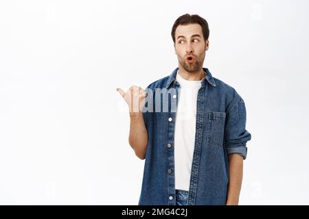 Excited man points left and looks amazed, say wow, impressed by smth, stands over white background Stock Photo
