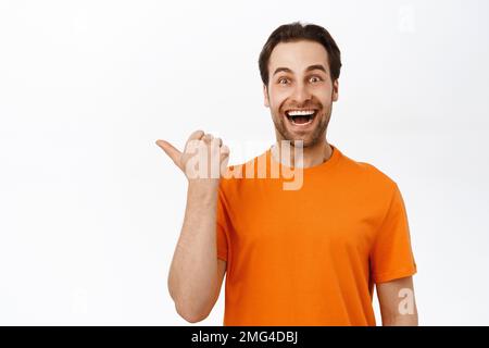 Close up of enthusiastic adult man pointing left, smiling amazed, showing advertisement aside, wearing orange t-shirt, white background Stock Photo