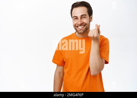 Portrait of cute guy, shows finger heart gesture and smiles, stands in orange tshirt against studio background Stock Photo