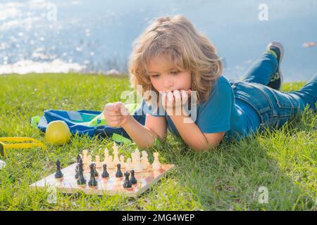 Concentrated child boy developing chess strategy, playing board game in backyard, laying on grass. Stock Photo