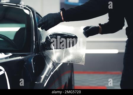 Unrecognisable man wearing black gloves leaning over stretching PPF Paint Protection Film over side car mirror. Professional car detailing process. Horizontal indoor shot. High quality photo Stock Photo