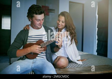 Hes special in so many ways. a young man playing the ukulele while sitting on bed with his girlfriend. Stock Photo