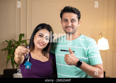 Smiling wife with husbnd showing house keys with thumbs up gesture by looking at camera at home - concept of insurance, new home purchase and Stock Photo