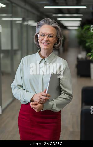 Attractive mature caucasian woman holding laptop while standing in office.  Stock Photo