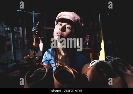 Beautiful Teenage Shop Girl Wearing Pink Leather Cap in Moody Shadowy Natural Window Light Surrounded by Shoes - Matte Stock Photo