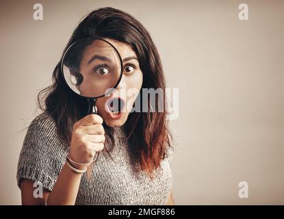 When you finally find what youve been stalking for. Studio portrait of a young woman looking shocked while using a magnifying glass against a brown Stock Photo