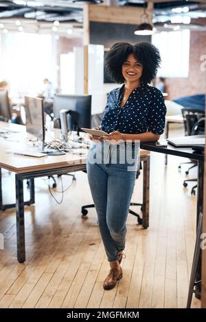 Her energy can be felt all over the office. Full length portrait of a cheerful young female designer holding a digital tablet while standing inside Stock Photo