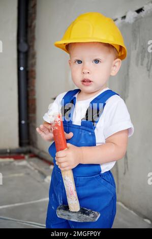 Adorable child construction worker holding hammer building repair tool and looking at camera. Cute kid wearing safety helmet and work overalls while posing at home during renovation. Stock Photo