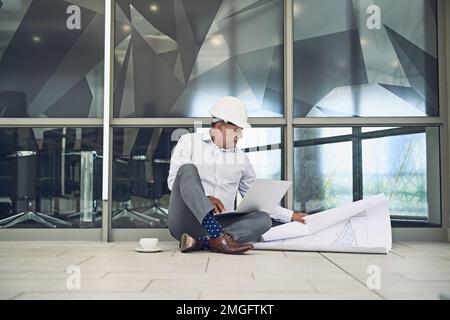 He will work wherever he is. a focused professional male architect sitting on the floor outside while working on a laptop. Stock Photo