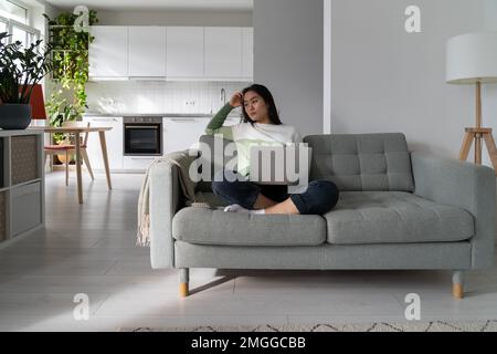 Thoughtful upset young Asian woman freelancer sitting on sofa with laptop looking aside Stock Photo