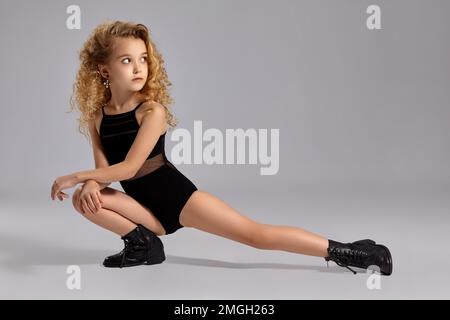 Beautiful little girl gymnast in a black sports swimsuit and boots on a gray background. Stock Photo