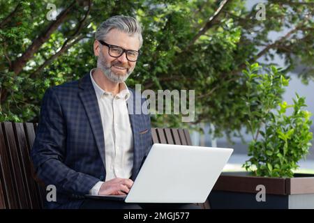 Portrait of senior businessman outdoors in park on bench, mature boss smiling and looking at camera, man in suit working on laptop remotely on sunny day. Stock Photo
