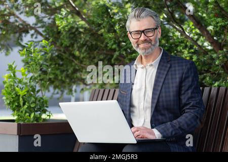 Portrait of senior businessman outdoors in park on bench, mature boss smiling and looking at camera, man in suit working on laptop remotely on sunny day. Stock Photo