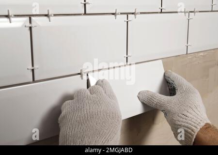 Male Worker Master Laying Ceramic Tiles on the Wall of Kitchen. Repairman Placing White Modern Tiles, Spreading Adhesive. Builder Installing Tile at R Stock Photo