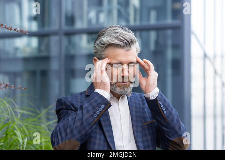 Sick and sad mature businessman outdoors sitting on bench on office building background, senior boss in suit having severe headache and dizziness, gray haired man wearing glasses outdoors. Stock Photo