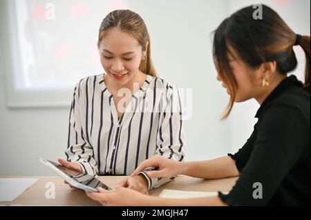 Attractive millennial Asian businesswoman using digital tablet, discussing work with her colleague in the office. Stock Photo