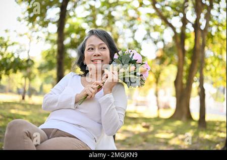 Charming and happy Asian 60s aged woman holding a beautiful flower bouquet while relaxing in the park. Stock Photo