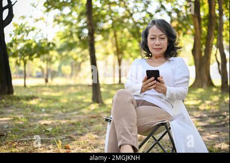Charming and happy Asian 60s aged woman in casual clothes using her smartphone, chatting with someone while relaxing in the park. Stock Photo
