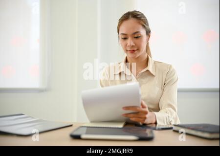 Focused millennial Asian businesswoman or female accountant examining sales and financial report at her desk, working in the office. Stock Photo
