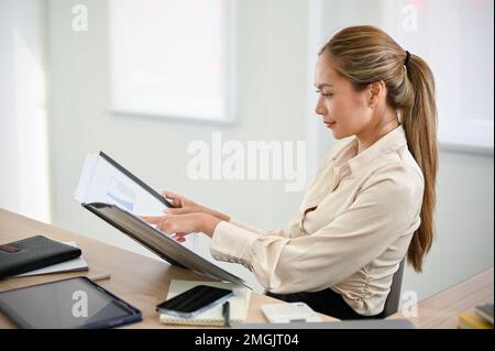 side view, Professional and attractive millennial Asian businesswoman or female manager examining business financial report on a folder binder at her Stock Photo