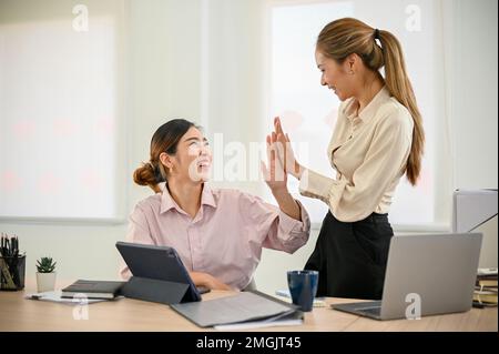 Two cheerful and happy millennial Asian female office workers give high fives to each other, celebrating their success, working together in the office Stock Photo