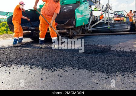 View on machine for laying asphalt, spreading layer of hot tarmac on prepared ground a few workers are using shovels to level it. Stock Photo